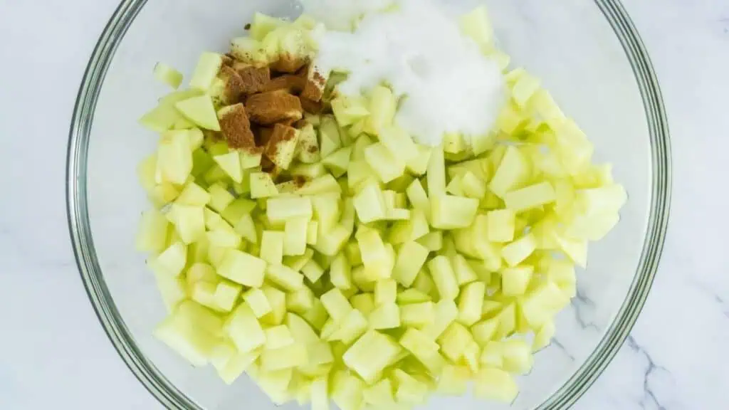 A glass bowl filled with diced green apples, granulated sugar, and brown spices on a white surface.