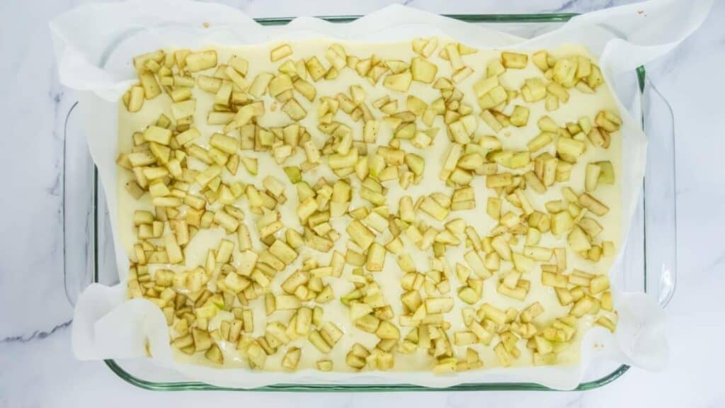 A parchment-lined glass baking dish filled with an unbaked dessert, topped with an even layer of chopped apples.