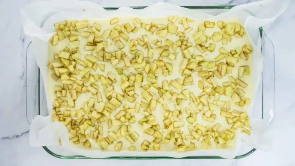 A parchment-lined glass baking dish filled with an unbaked dessert, topped with an even layer of chopped apples.