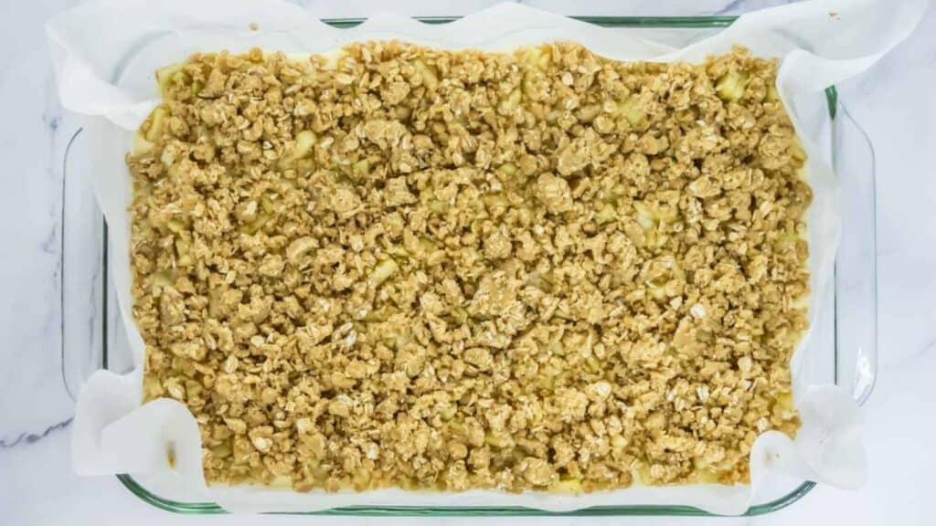 A glass baking dish lined with parchment paper contains an unbaked oat crumble mixture, spread evenly across the top.