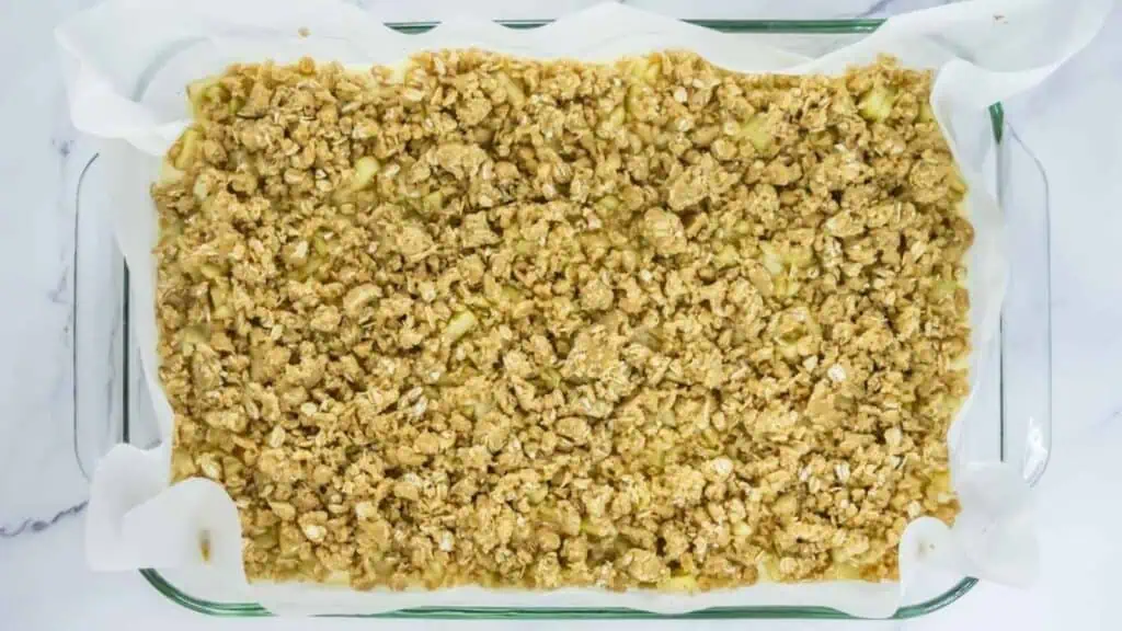 A glass baking dish lined with parchment paper contains an unbaked oat crumble mixture, spread evenly across the top.