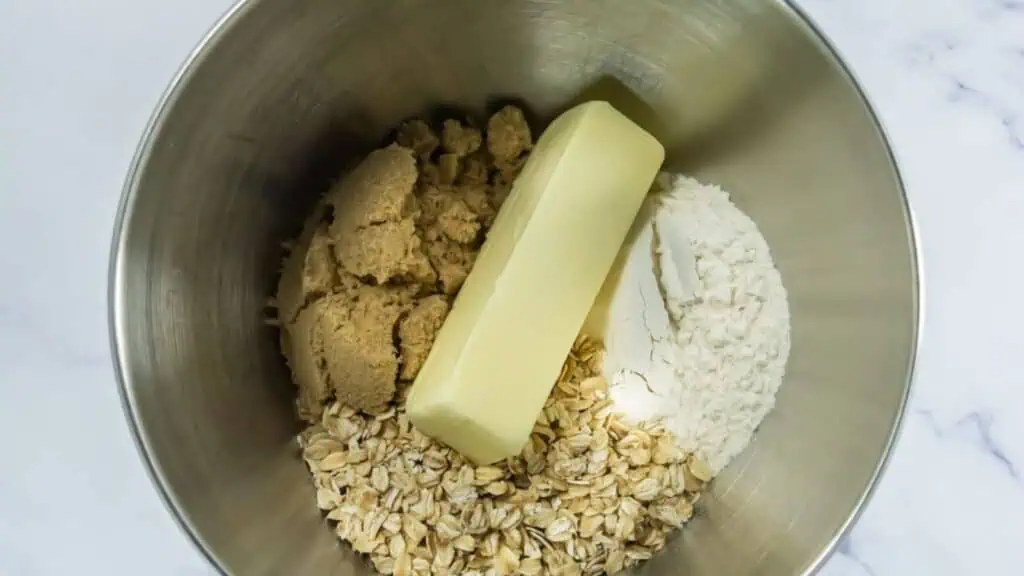 A metal mixing bowl containing brown sugar, a stick of butter, rolled oats, and flour.