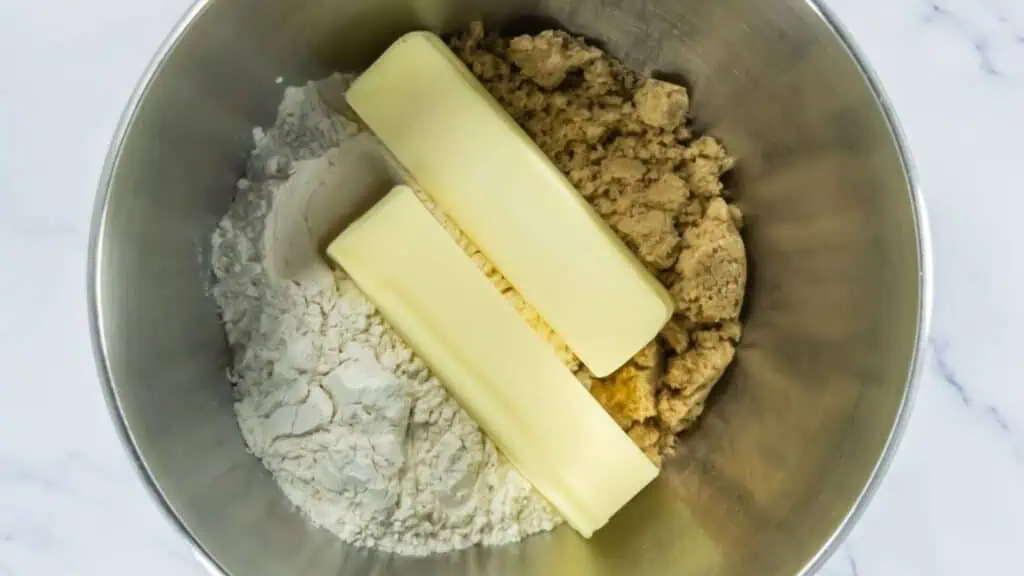 A metal mixing bowl contains flour, brown sugar, and two sticks of butter, ready to be mixed for baking.