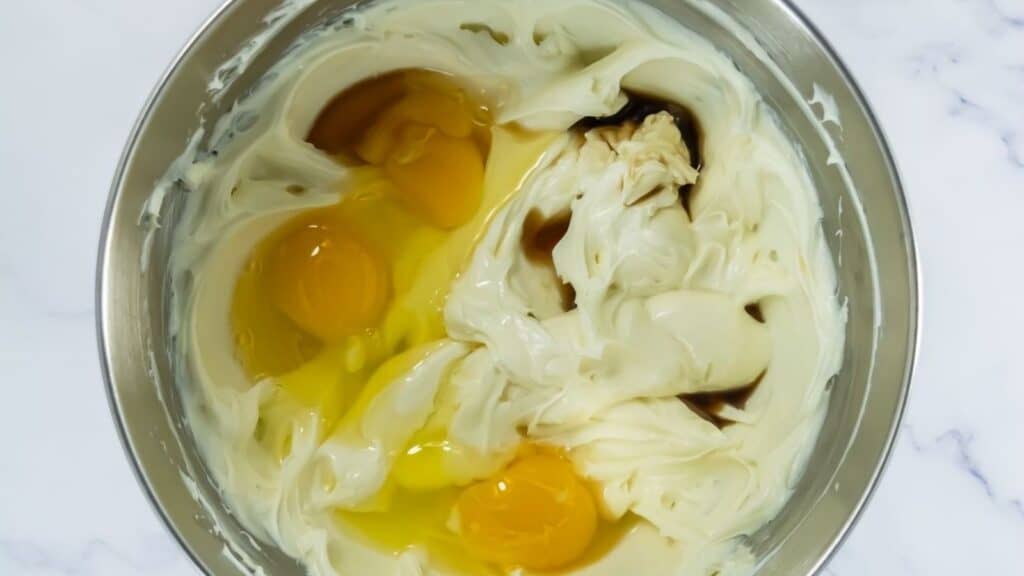 Three raw eggs, vanilla extract, and creamed mixture are in a metal mixing bowl, viewed from above on a white countertop.