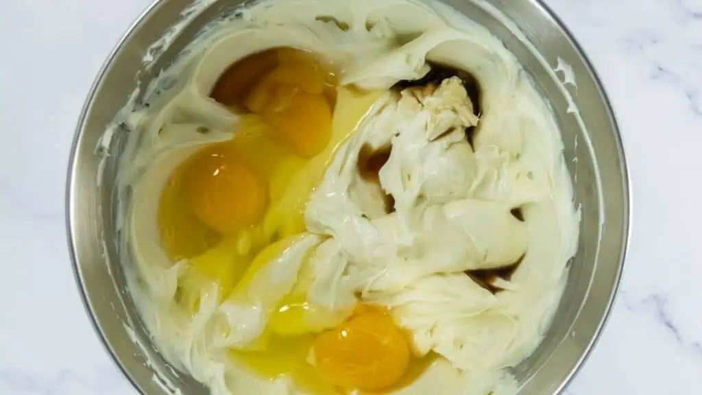 Three raw eggs, vanilla extract, and creamed mixture are in a metal mixing bowl, viewed from above on a white countertop.