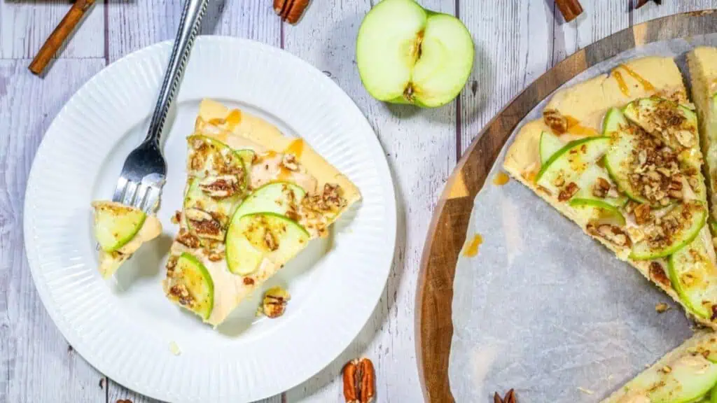 A slice of apple and pecan tart is served on a white plate with a fork, next to the remaining tart on a wooden board and a sliced green apple.