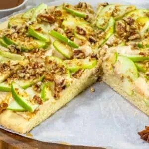 A dessert pizza topped with sliced green apples, chopped pecans, caramel drizzle, and cream cheese frosting, with one slice missing.