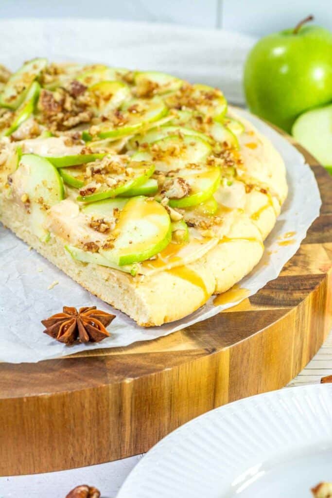 A dessert pizza topped with sliced green apples, chopped nuts, and caramel drizzle sits on parchment paper on a wooden board.