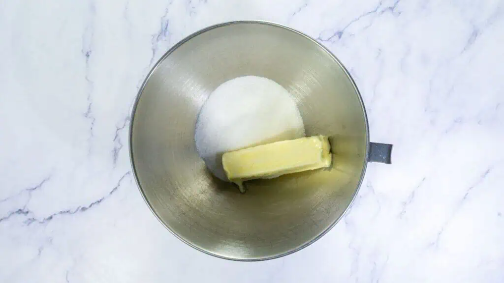 A metal mixing bowl containing a stick of butter and granulated sugar on a white marble surface.