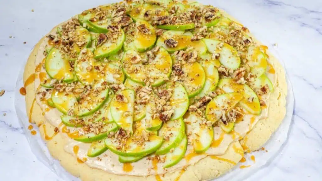 A dessert pizza topped with sliced green apples, chopped nuts, and caramel sauce on a baked crust.