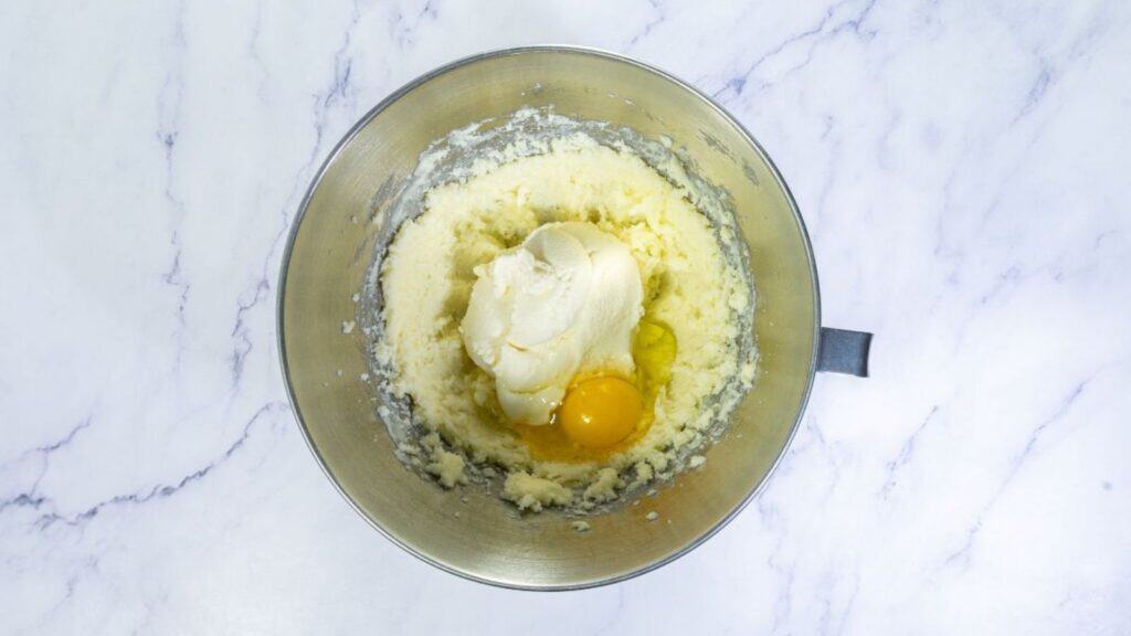 A mixing bowl containing creamed butter and sugar, with a dollop of cream cheese and an egg on top, on a white marble surface.