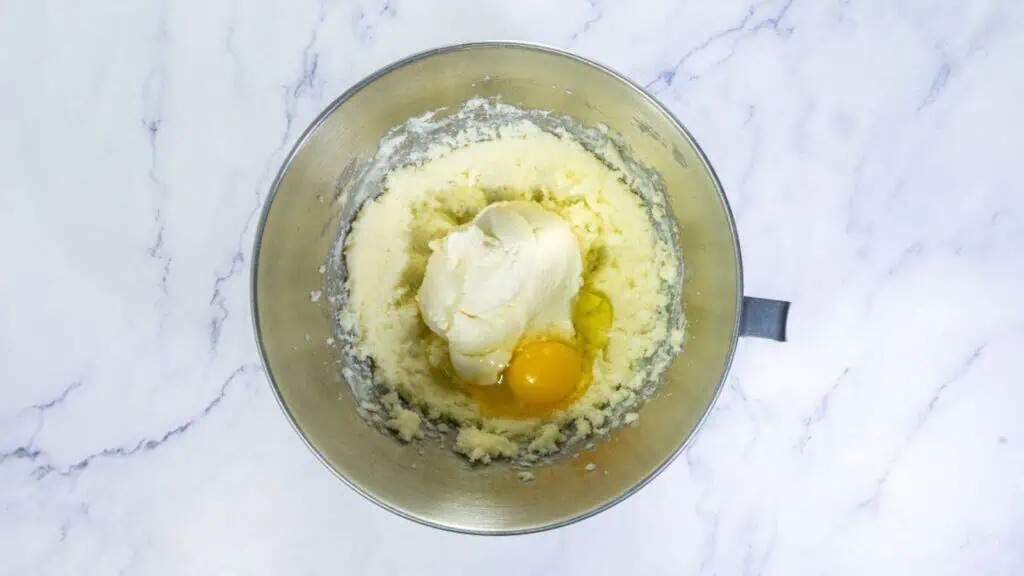 A mixing bowl containing creamed butter and sugar, with a dollop of cream cheese and an egg on top, on a white marble surface.
