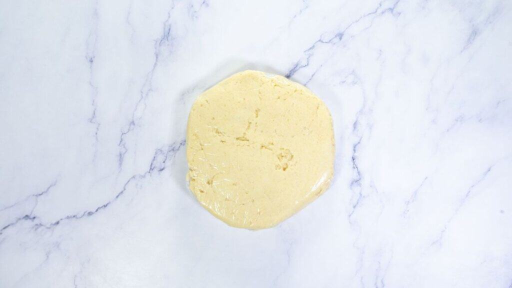 A round disk of dough wrapped in plastic wrap sits on a white marble surface.
