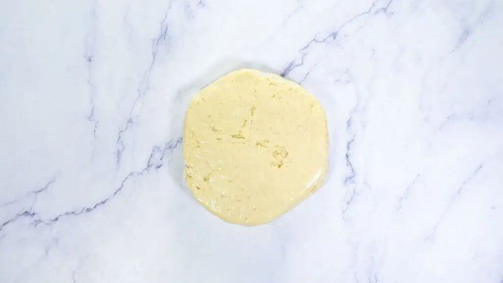 A round disk of dough wrapped in plastic wrap sits on a white marble surface.