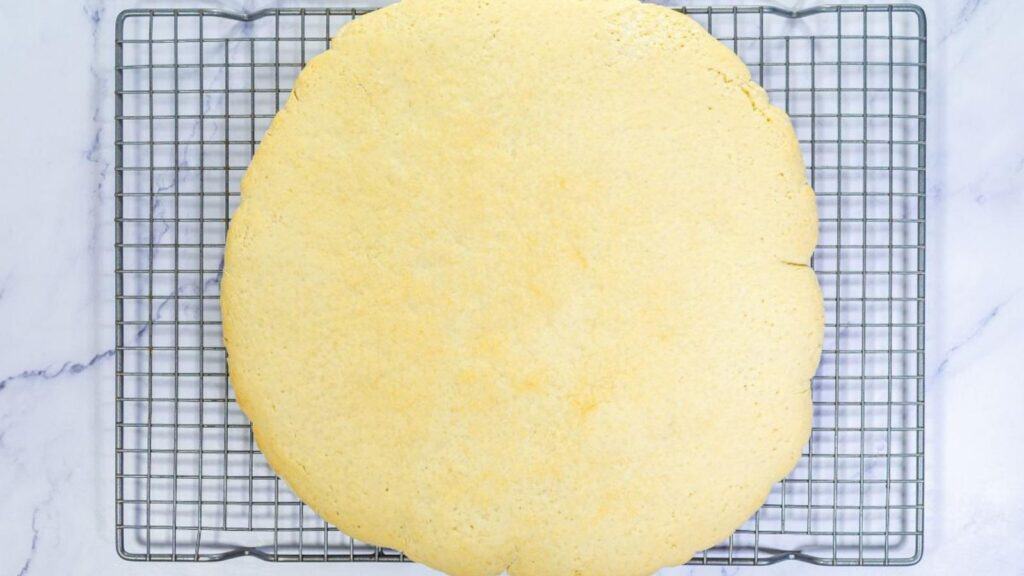 A large, round, plain baked cookie resting on a metal cooling rack over a white marble surface.