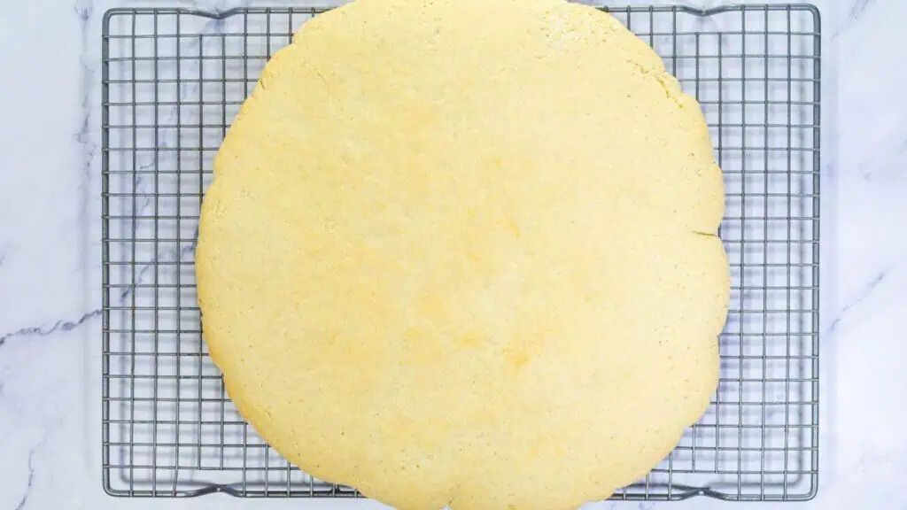 A large, round, plain baked cookie resting on a metal cooling rack over a white marble surface.