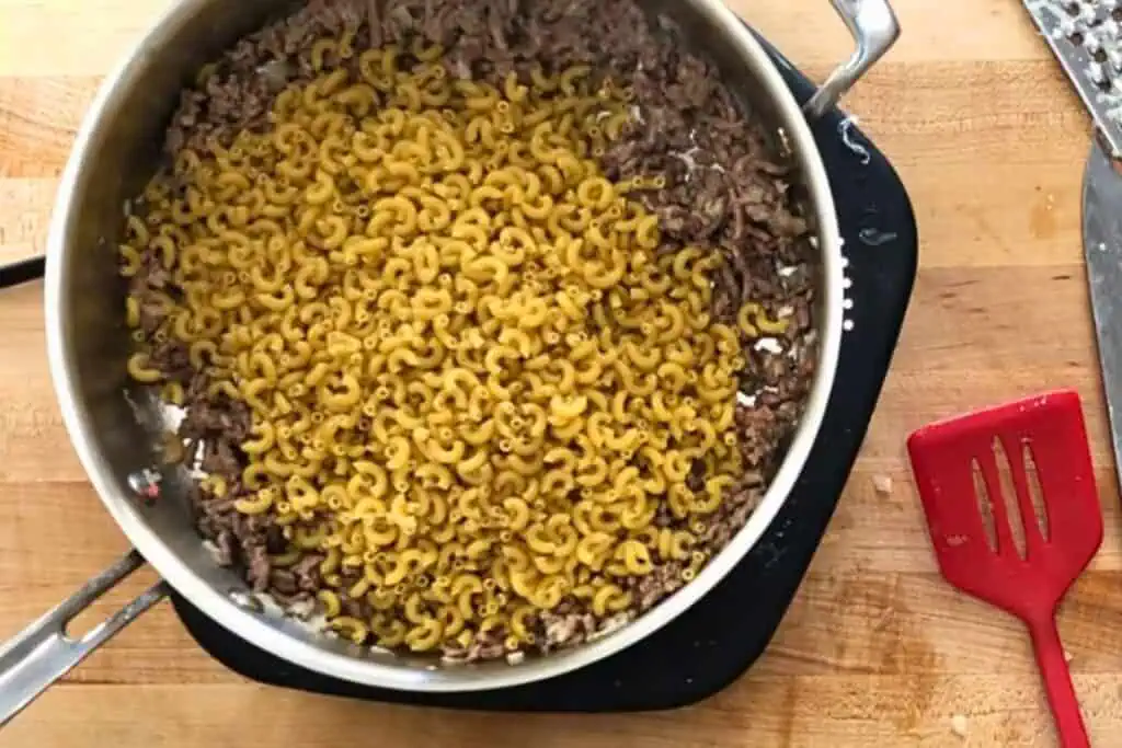 A pot on a wooden counter contains cooked ground beef and uncooked elbow macaroni. A red spatula is next to the pot.