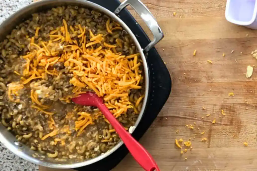 A pot of macaroni and ground beef topped with shredded cheddar cheese sits on a stovetop burner, with a red spatula and cheese scattered on a wooden counter nearby.