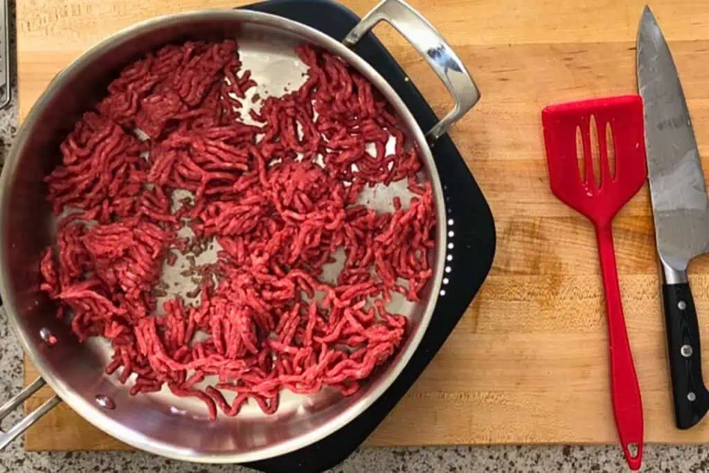 Ground beef being browned in a stainless steel pan on a stovetop, with a red spatula and a knife on a wooden cutting board nearby.