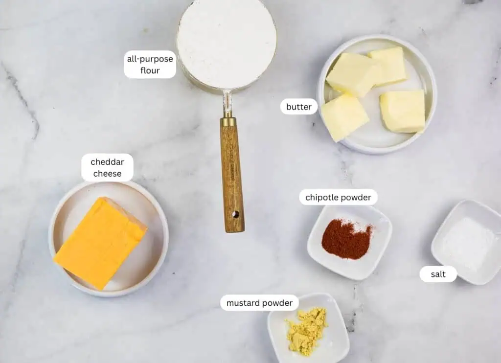 Ingredients on a marble surface: all-purpose flour, butter, cheddar cheese, chipotle powder, mustard powder, and salt, each labeled in separate dishes.