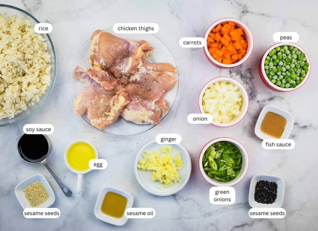 Ingredients for a chicken fried rice recipe displayed on a marble surface, including chicken thighs, rice, vegetables, sauces, sesame seeds, oil, egg, and seasonings.