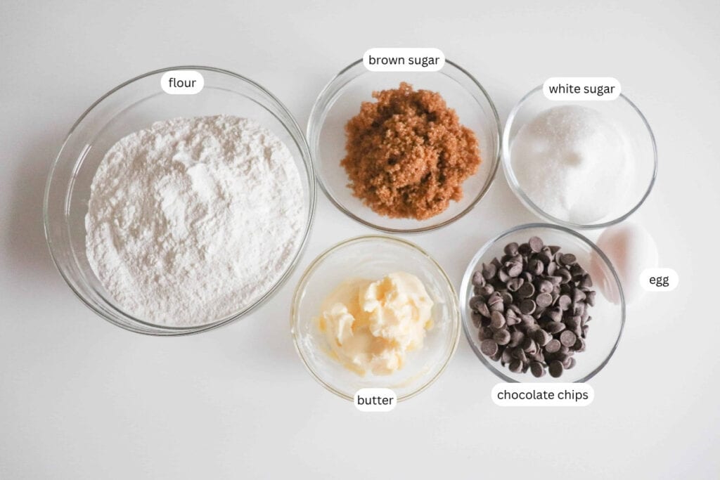 Five bowls containing labeled ingredients: flour, brown sugar, white sugar, butter, and chocolate chips, plus one labeled egg, arranged on a white surface.