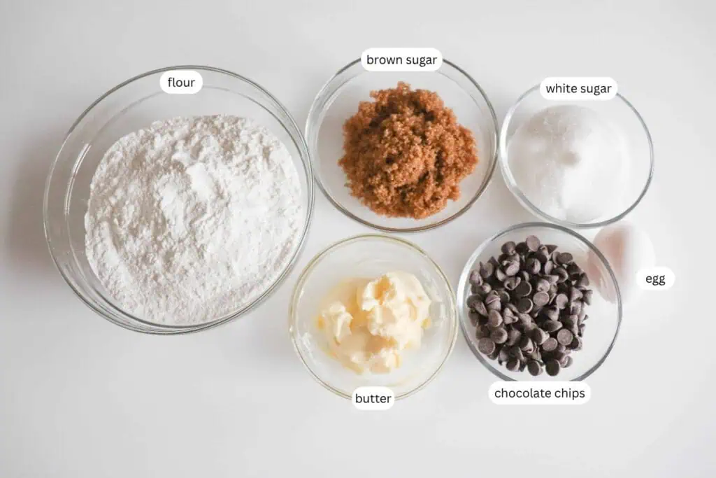 Five bowls containing labeled ingredients: flour, brown sugar, white sugar, butter, and chocolate chips, plus one labeled egg, arranged on a white surface.