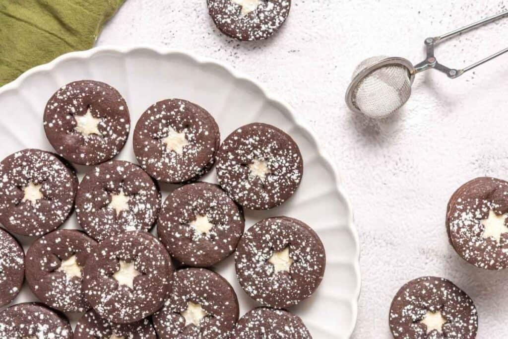 A plate of round chocolate cookies with powdered sugar and a star-shaped center, next to a tea strainer with sugar on a light surface.