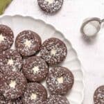 A plate of round chocolate cookies with powdered sugar and a star-shaped center, next to a tea strainer with sugar on a light surface.