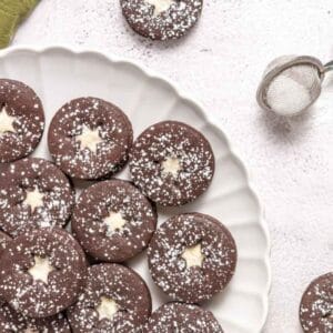 A plate of round chocolate cookies with powdered sugar and a star-shaped center, next to a tea strainer with sugar on a light surface.