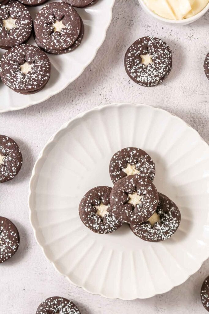 Round chocolate sandwich cookies with star-shaped cutouts and powdered sugar dusting arranged on white plates, with more cookies and a small bowl nearby.