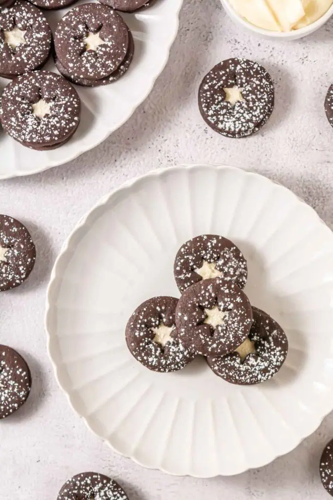 Round chocolate sandwich cookies with star-shaped cutouts and powdered sugar dusting arranged on white plates, with more cookies and a small bowl nearby.