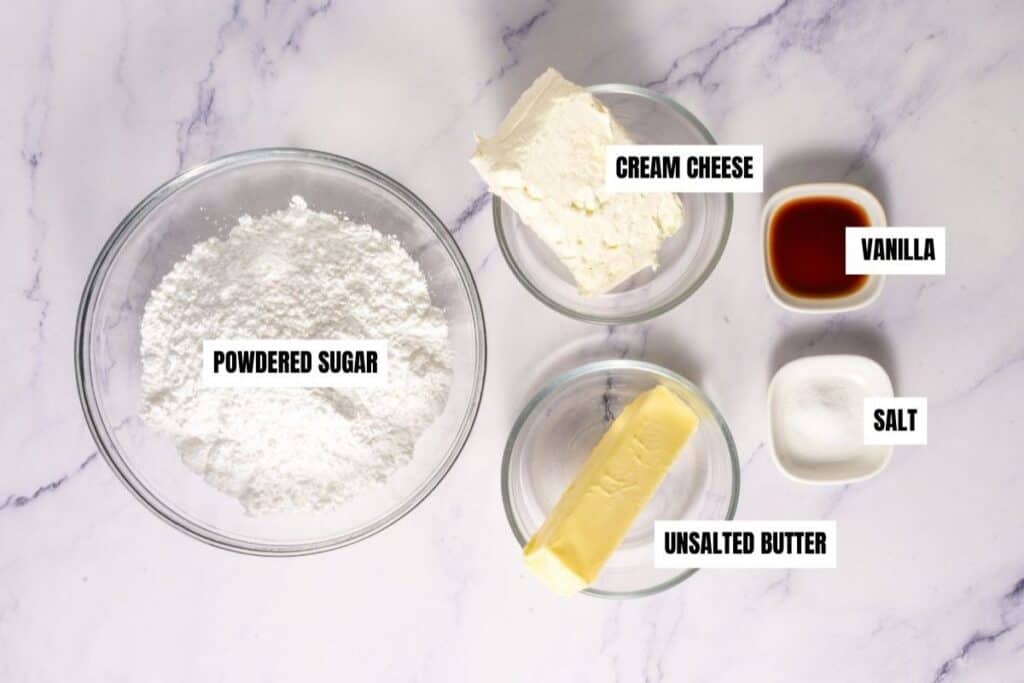 Five labeled bowls containing powdered sugar, cream cheese, vanilla, salt, and unsalted butter are arranged on a white marble surface.