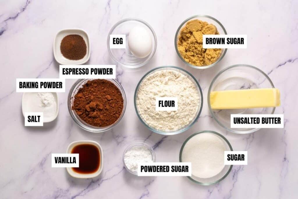 Bowls containing labeled baking ingredients: espresso powder, egg, brown sugar, baking powder, salt, cocoa powder, flour, unsalted butter, vanilla, powdered sugar, and sugar on a marble surface.
