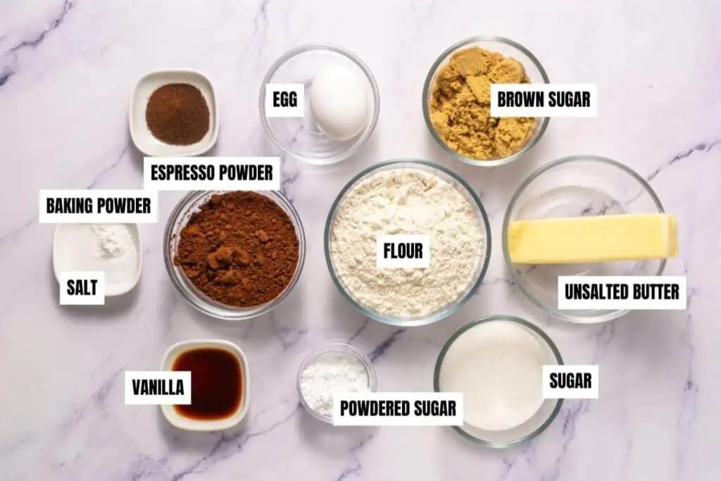 Bowls containing labeled baking ingredients: espresso powder, egg, brown sugar, baking powder, salt, cocoa powder, flour, unsalted butter, vanilla, powdered sugar, and sugar on a marble surface.