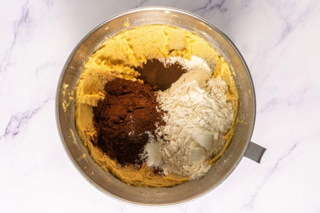 A metal mixing bowl with creamed batter, cocoa powder, flour, and spices on top, ready to be mixed together, on a light marble surface.
