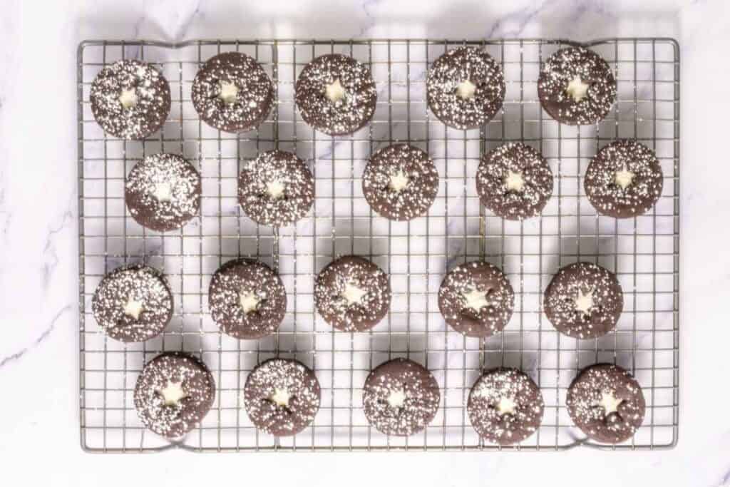 A cooling rack with twenty chocolate cookies topped with powdered sugar and a star-shaped white center, arranged in neat rows on a white surface.