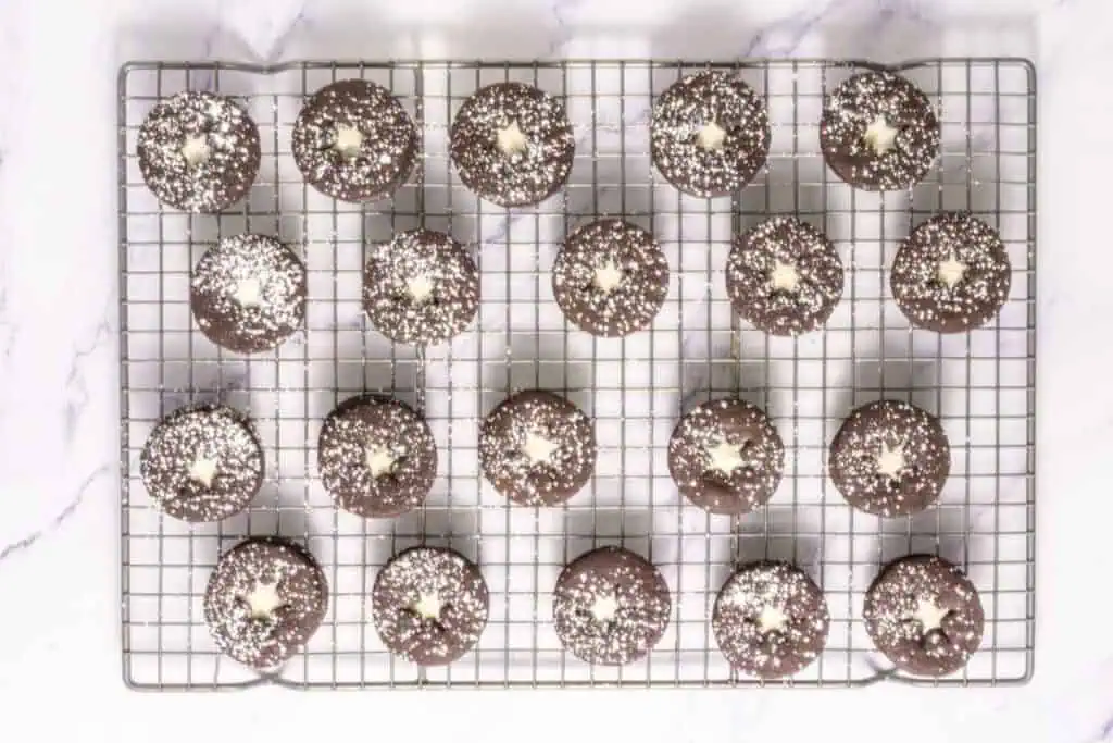 A cooling rack with twenty chocolate cookies topped with powdered sugar and a star-shaped white center, arranged in neat rows on a white surface.