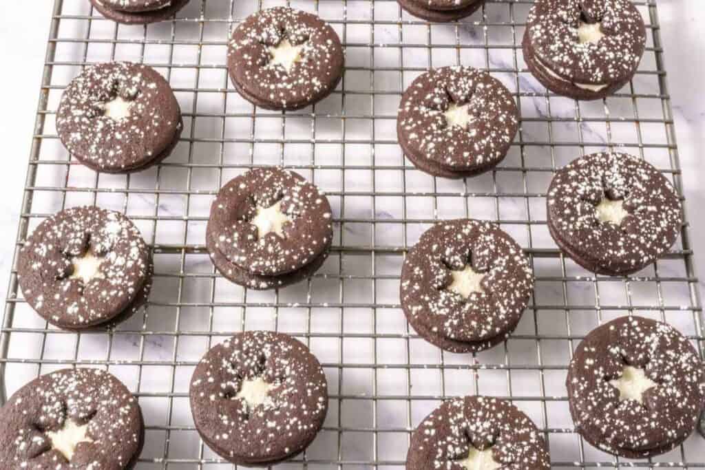 Chocolate sandwich cookies with star-shaped cutouts and powdered sugar on top, arranged in rows on a cooling rack.