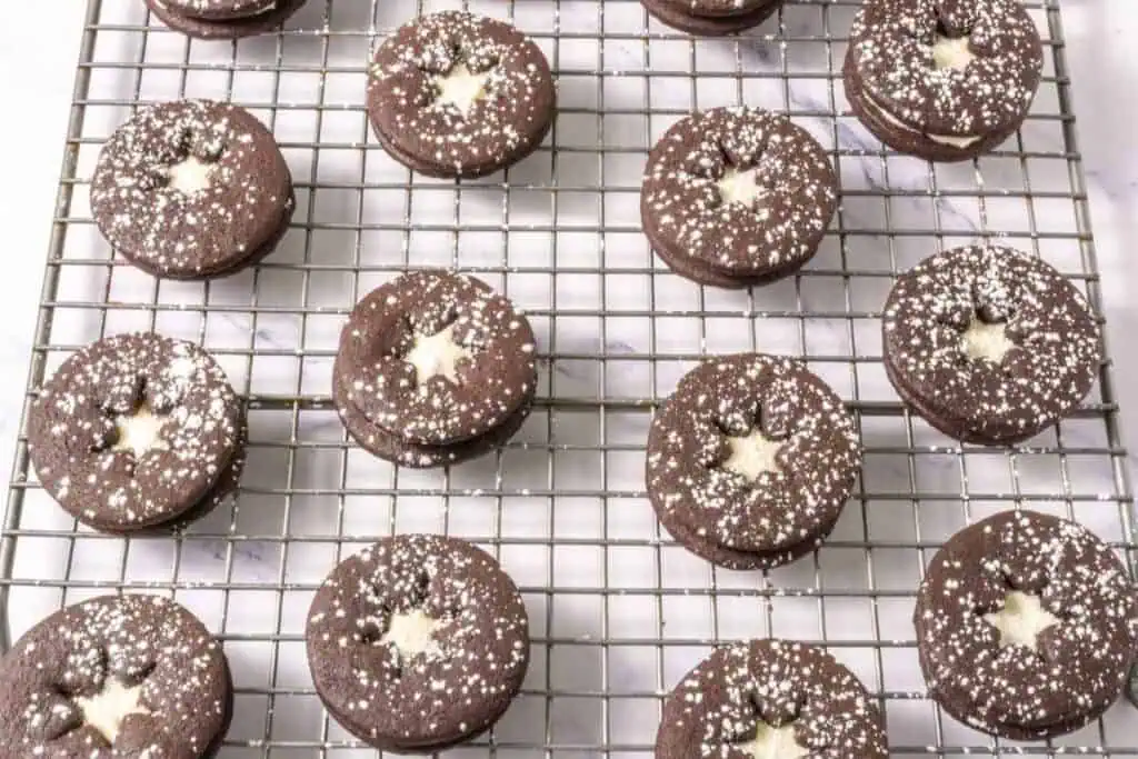 Chocolate sandwich cookies with star-shaped cutouts and powdered sugar on top, arranged in rows on a cooling rack.