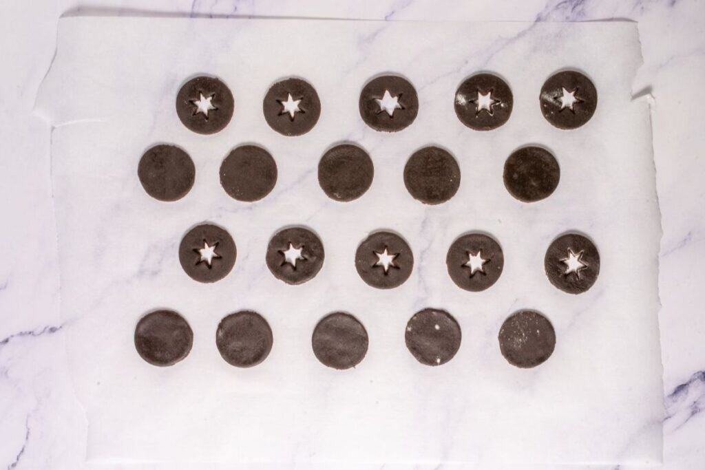 Round chocolate cookie dough pieces on parchment paper, some with star-shaped cutouts in the center, arranged in rows on a light marble surface.