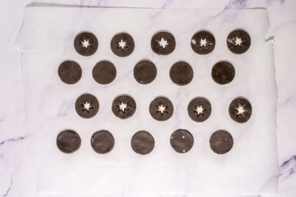Round chocolate cookie dough pieces on parchment paper, some with star-shaped cutouts in the center, arranged in rows on a light marble surface.