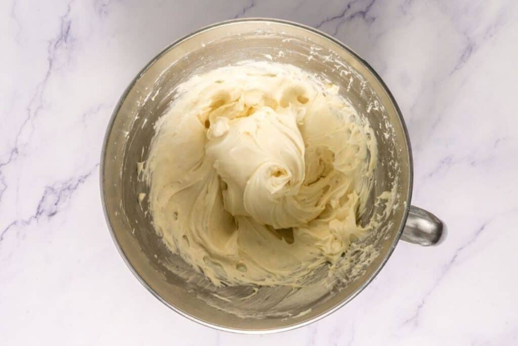 A metal mixing bowl filled with thick, creamy frosting sits on a white marble surface.
