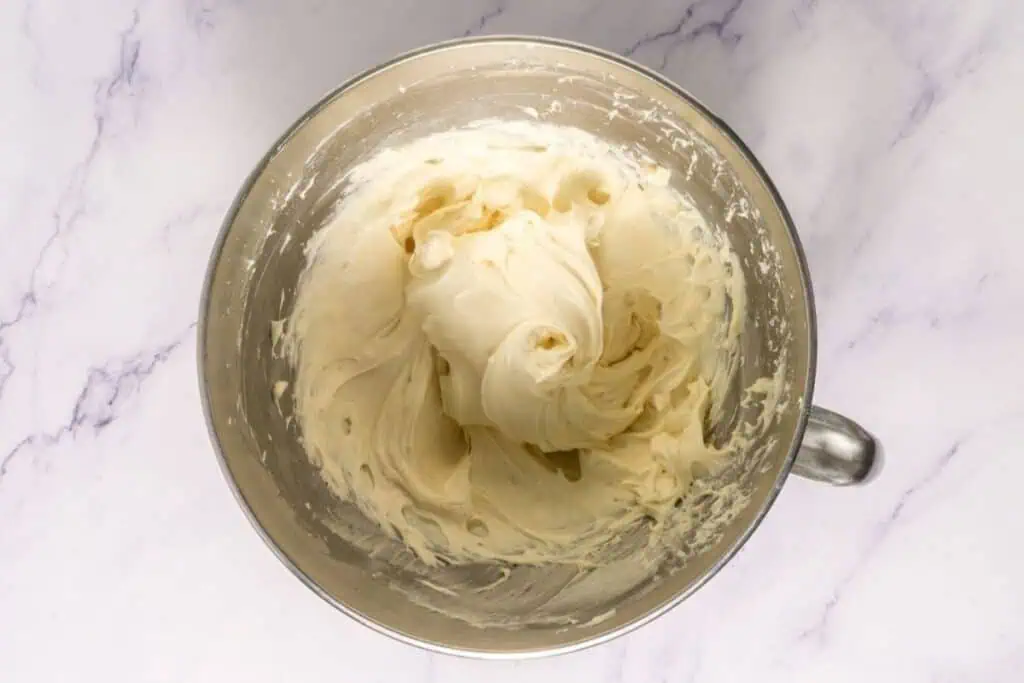 A metal mixing bowl filled with thick, creamy frosting sits on a white marble surface.