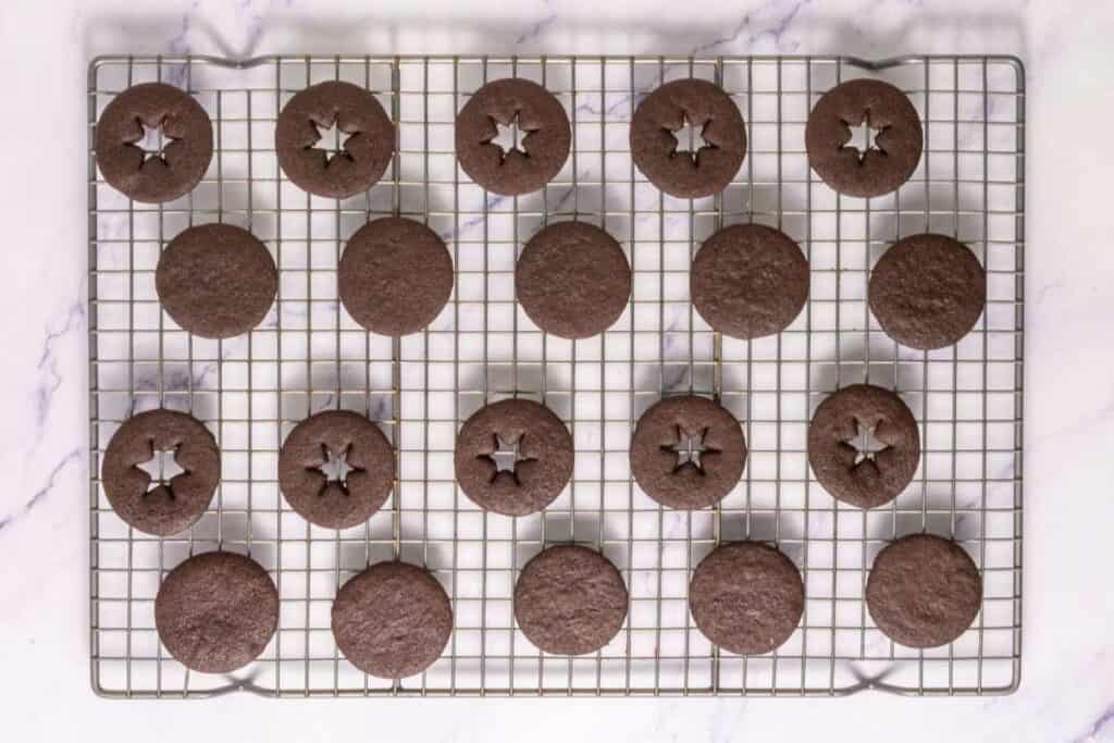 A cooling rack with 20 round chocolate cookies, some with star-shaped cutouts in the center, on a white marble surface.