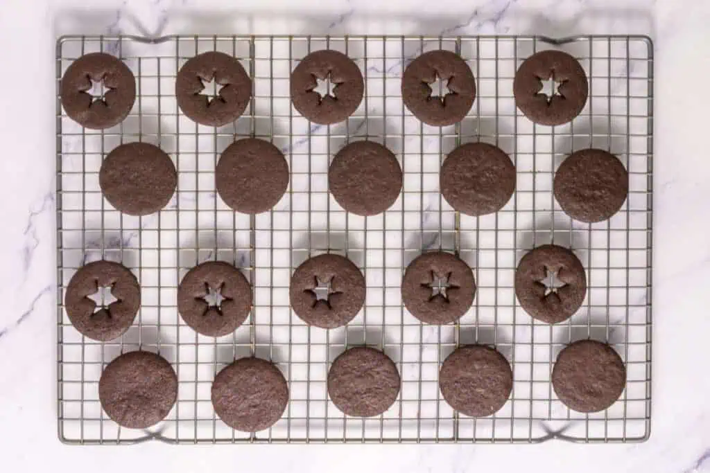A cooling rack with 20 round chocolate cookies, some with star-shaped cutouts in the center, on a white marble surface.
