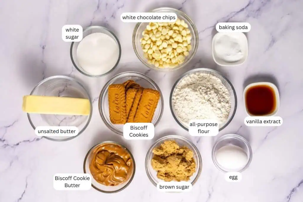 Various baking ingredients, including unsalted butter, white sugar, Biscoff cookies, Biscoff cookie butter, white chocolate chips, brown sugar, flour, baking soda, vanilla extract, and an egg.