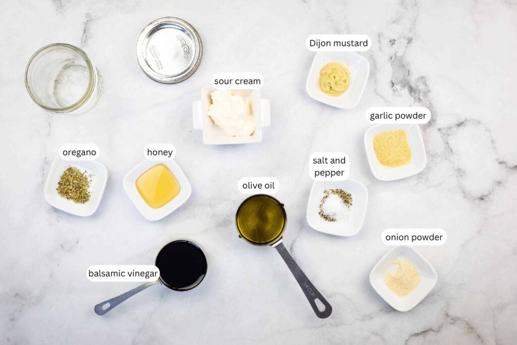 Ingredients for a dressing are arranged on a marble surface, each labeled: sour cream, Dijon mustard, garlic powder, onion powder, salt and pepper, olive oil, balsamic vinegar, honey, oregano, and an empty jar.