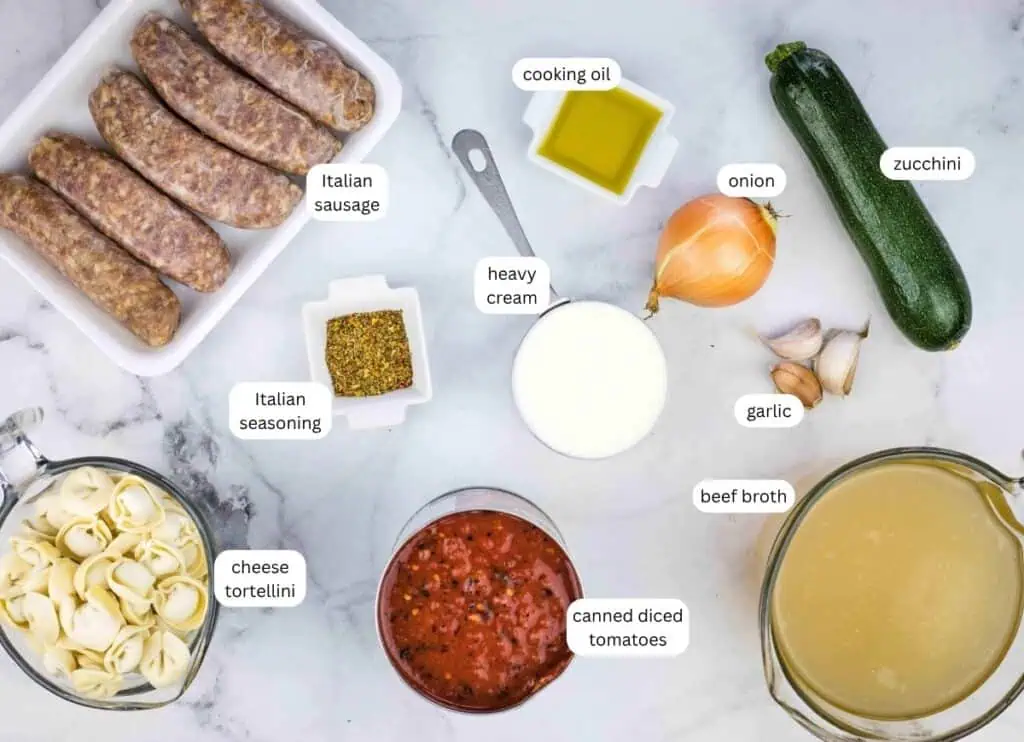 Top-down view of recipe ingredients: Italian sausage, cheese tortellini, Italian seasoning, canned diced tomatoes, beef broth, heavy cream, cooking oil, onion, garlic, and zucchini.