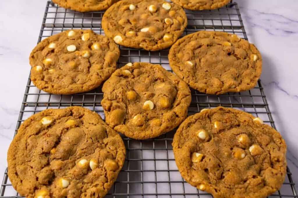 Nine large cookies with white chocolate chips cooling on a metal wire rack on a marble surface.